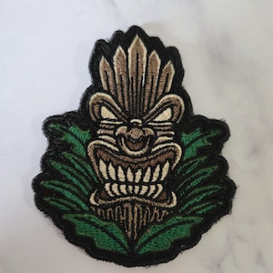 May include: A brown and green embroidered patch featuring a tiki head with a menacing expression. The tiki head is surrounded by green leaves.
