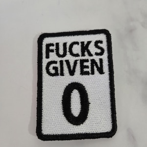 May include: A rectangular embroidered patch with a white background and a thick black border. The patch features the black embroidered text "FUCKS GIVEN" in two lines at the top, followed by a large black embroidered number "0" centred below.