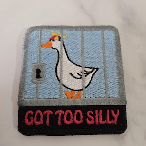 Puede incluir: Parche bordado con un ganso blanco con un sombrero pequeño, dentro de una jaula azul con barras grises. El parche tiene un borde negro y las palabras "GOT TOO SILLY" en rojo.