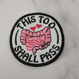 May include: Embroidered patch with the text "THIS TOO SHALL PASS" in a circular design. The patch features a pink illustration of intestines with two red hearts above. The border is black.