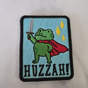 Op de afbeelding: Een rechthoekige geborduurde patch met een lichtblauwe achtergrond en een zwarte rand. Het toont een groene kikker met een rode cape, die een zilveren zwaard vasthoudt, met gele ruitvormen en de zwarte tekst HUZZAH! onderaan gedrukt.