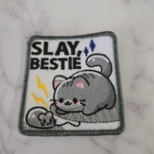 Könnte beinhalten: Gestickter Aufnäher mit einem Cartoon-Katzen- und Maus-Design. Die graue Katze hat rosa Details und liegt neben einer grauen Maus. Der Aufnäher trägt den Schriftzug "SLAY, BESTIE" in schwarzer Schrift, mit gelben Blitzen und blauen Diamanten.