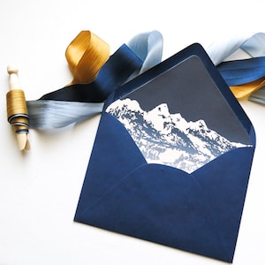 May include: A navy blue envelope with a white mountain range design printed on the inside flap. The envelope is decorated with blue and gold ribbon.