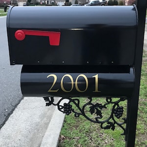 May include: Black mailbox with a red flag and gold lettering reading "2001". The mailbox is mounted on a black post with decorative grape vine accents. The mailbox is located on a residential street.