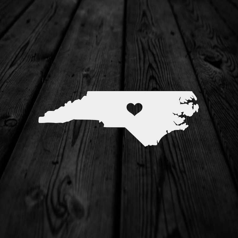 May include: A white outline of the state of North Carolina with a black heart shape in the center. The image is set against a dark, textured wooden background, creating a contrast between the bright white and the dark wood.