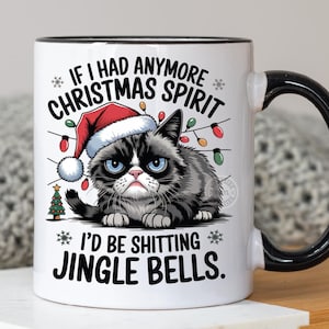 Peut inclure: Mug en céramique blanche avec intérieur et anse noirs. Le mug présente un chat grincheux portant un bonnet de Père Noël, entouré de guirlandes lumineuses et du texte : « Si j'avais plus d'esprit de Noël, je chierais des grelots. »
