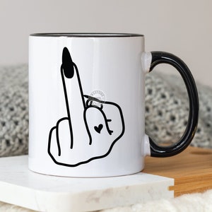 Puede incluir: Taza de cerámica blanca con borde y asa negros. La taza presenta una ilustración en blanco y negro de una mano haciendo el dedo medio con un anillo en el dedo medio y un pequeño tatuaje de corazón en el dedo índice.