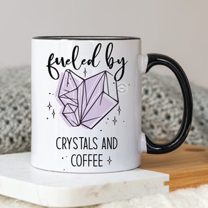 May include: White ceramic mug with a black handle and rim. The mug features the text "fueled by crystals and coffee" with a light purple crystal graphic. The mug is on a white marble surface.