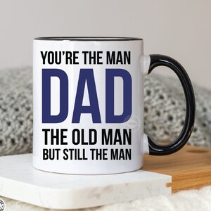 May include: White ceramic mug with a black rim and handle. The mug has a blue and black design that reads "You're the man Dad The old man but still the man."