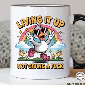 May include: White ceramic mug with a black handle and rim, featuring a cartoon duck wearing heart-shaped sunglasses and pink clogs. The mug has the text "LIVING IT UP" and "NOT GIVING A FUCK" with a rainbow and flowers.