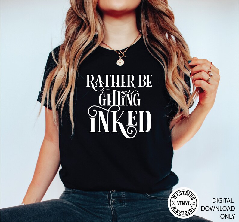 May include: A black t-shirt with white text that reads "Rather Be Getting Inked". The shirt is being worn by a person with long blonde hair. The image also includes a logo for Westside Vinyl.