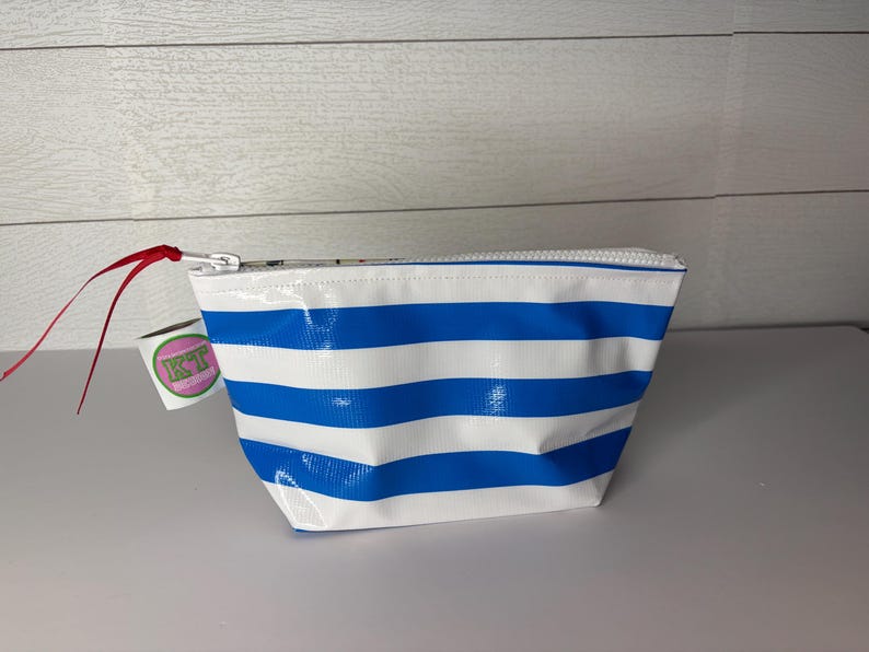 May include: A white and blue striped cosmetic bag with a zipper closure. The bag has a red ribbon attached to the zipper pull. A small tag with the letters "KT" is attached to the bag.