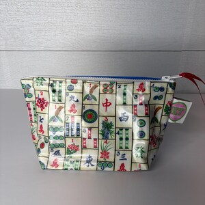 May include: A small, rectangular cosmetic bag with a blue zipper and a red pull tab. The bag is covered in a repeating pattern of colorful Mahjong tiles. A small tag with text is attached to the side.