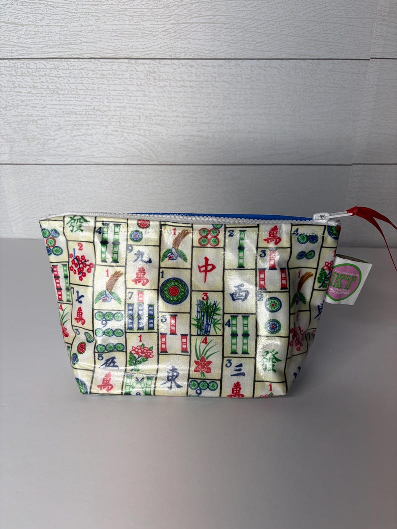 May include: A small, rectangular cosmetic bag with a blue zipper and a red pull tab. The bag is covered in a repeating pattern of colorful Mahjong tiles. A small tag with text is attached to the side.
