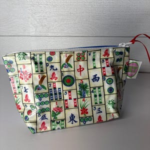 May include: A small, rectangular pouch with a zipper closure. The pouch is covered in a pattern of colorful Mahjong tiles on a cream background. The zipper is blue, and a red string is attached to the zipper pull.