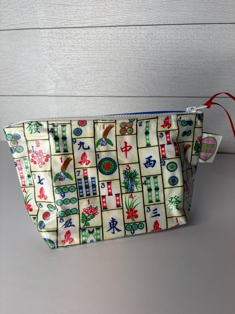 May include: A small, rectangular pouch with a zipper closure. The pouch is covered in a pattern of colorful Mahjong tiles on a cream background. The zipper is blue, and a red string is attached to the zipper pull.