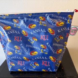 May include: A blue zippered pouch featuring a repeating pattern of the University of Kansas Jayhawks logo and text. The design includes the words "KANSAS", "JAYHAWKS", and "KU". A red cord is attached to the zipper pull. A small tag with "KT" is attached.