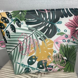 May include: A white zippered pouch with a tropical leaf print in shades of green, pink, and yellow. The pouch has a white background with a variety of leaf shapes and patterns.