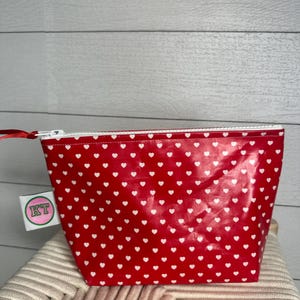 May include: Red zippered pouch with a white heart pattern. The pouch has a white zipper and a white tag with the letters "KT" in pink.