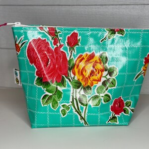 May include: A turquoise zippered pouch with a floral pattern featuring red and yellow roses. The pouch has a white zipper and a small loop on the side. The background is a grid pattern.