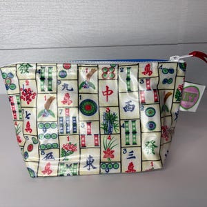 May include: A small, rectangular pouch with a zipper closure. The pouch is covered in a pattern of colorful Mahjong tiles. The zipper is white with a red pull, and the interior is blue. A small tag with a logo is attached.