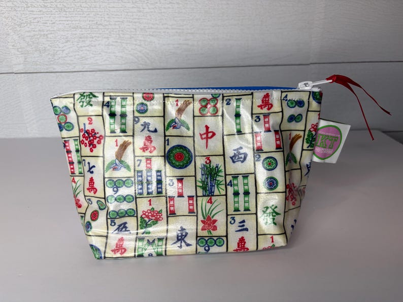 May include: A small, rectangular pouch with a zipper closure. The pouch is covered in a pattern of colorful Mahjong tiles. The zipper is white with a red pull, and the interior is blue. A small tag with a logo is attached.