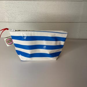 May include: A small, white and blue striped cosmetic bag with a zipper closure. The bag has a rectangular shape and a red pull tab. A small tag with the letters "KT" is attached to the zipper.