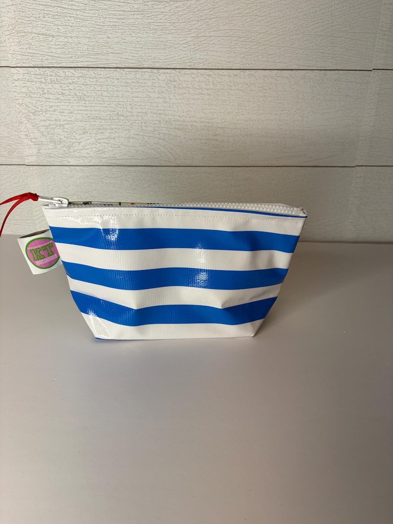 May include: A small, white and blue striped cosmetic bag with a zipper closure. The bag has a rectangular shape and a red pull tab. A small tag with the letters "KT" is attached to the zipper.