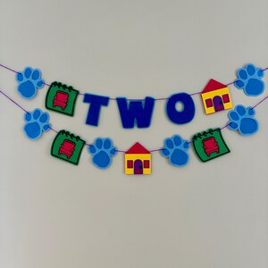 May include: A blue and multi-colored banner with the word "TWO" in large blue letters. The banner also features blue paw prints, red houses with yellow accents, and green notepads with red chairs. The banner is strung on a purple string.