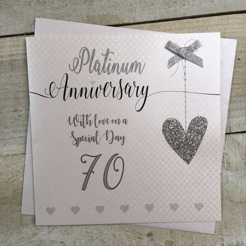 Puede incluir: Una tarjeta de brillo plateado con un coraz&oacute;n y un lazo. La tarjeta dice "Aniversario de Platino" y "Con amor en un d&iacute;a especial 70".