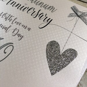Puede incluir: Una tarjeta blanca con detalles de purpurina plateada. La tarjeta dice "Aniversario de Platino con amor en un d&iacute;a especial".
