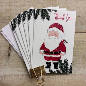 May include: A stack of white greeting cards featuring a watercolor illustration of Santa Claus wearing a red suit and hat with a small bird on his shoulder. The cards include green pine branch accents and the text Thank You in red script.