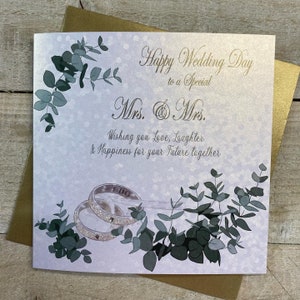 K&ouml;nnte beinhalten: Eine wei&szlig;e Karte mit goldfarbenem Rand und einem goldenen Glitzerdesign. Die Karte sagt "Happy Wedding Day to a Special Mrs. & Mrs. Wishing you Love, Laughter & Happiness for your Future together." Es gibt zwei silberne Ringe mit den Worten "I Do" darauf.