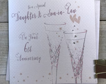 May include: A card with a pink background and silver glitter accents. It says "To a special Daughter & Son-in-Law On Your 6th Anniversary" with two champagne glasses and two hearts.