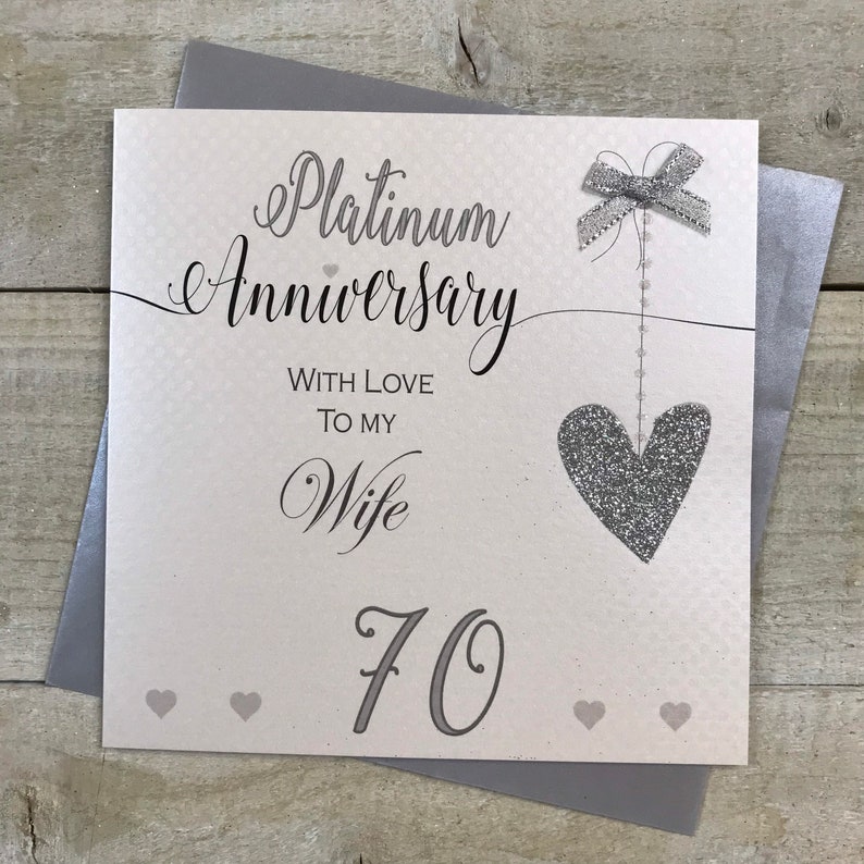 Puede incluir: Una tarjeta blanca con un coraz&oacute;n y un lazo de purpurina plateada. La tarjeta dice "Aniversario de platino" con amor para mi esposa 70.