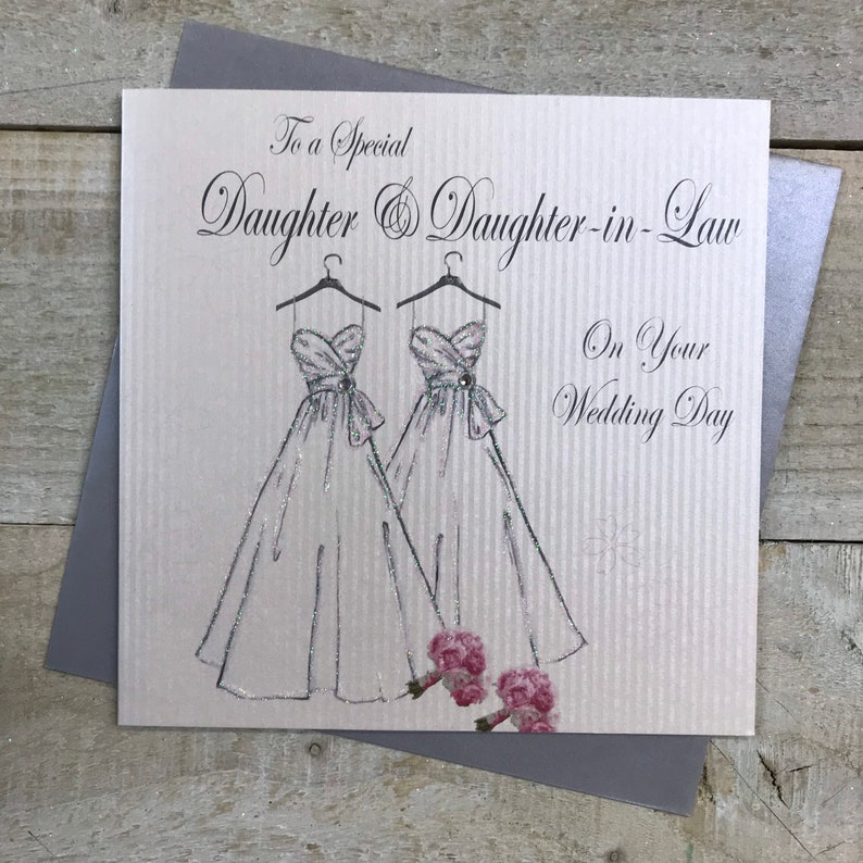 May include: A wedding card with a silver glitter background and two white wedding dresses with pink flowers. The card reads "To a special Daughter & Daughter-in-Law On Your Wedding Day".