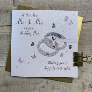K&ouml;nnte beinhalten: Eine wei&szlig;e Karte mit einem silbernen Glitzerdesign aus zwei Eheringen mit den Worten "I DO" auf einem der Ringe. Die Karte tr&auml;gt den Text "To the New Mrs. & Mrs. on your Wedding Day" und "Wishing you a happily ever after".