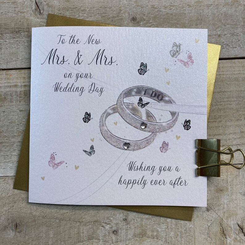 K&ouml;nnte beinhalten: Eine wei&szlig;e Karte mit einem silbernen Glitzerdesign aus zwei Eheringen mit den Worten "I DO" auf einem der Ringe. Die Karte tr&auml;gt den Text "To the New Mrs. & Mrs. on your Wedding Day" und "Wishing you a happily ever after".