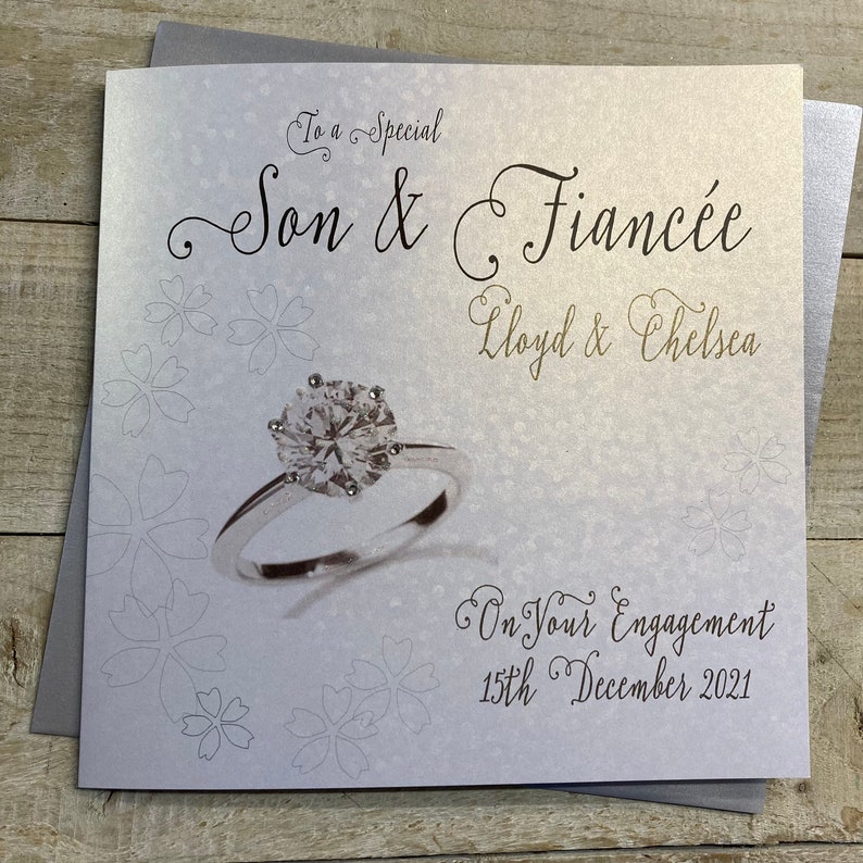 K&ouml;nnte beinhalten: Eine wei&szlig;e Karte mit silbernem Schimmer. Die Karte zeigt eine Diamant-Ring-Illustration und den Text "To a Special Son & Fianc&eacute;e Lloyd & Chelsea On Your Engagement 15th December 2021".
