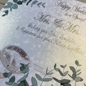 K&ouml;nnte beinhalten: Eine Hochzeitskarte mit wei&szlig;em Hintergrund und goldener Schrift. Die Karte sagt "Happy Wedding to a Special Mrs. & Mrs. Wishing you Love, Laughter & Happiness for your Future together." Es gibt zwei goldene Ringe, auf einem steht "I Do". Die Karte ist mit gr&uuml;nen Bl&auml;ttern verziert.