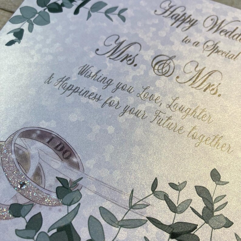 K&ouml;nnte beinhalten: Eine Hochzeitskarte mit wei&szlig;em Hintergrund und goldener Schrift. Die Karte sagt "Happy Wedding to a Special Mrs. & Mrs. Wishing you Love, Laughter & Happiness for your Future together." Es gibt zwei goldene Ringe, auf einem steht "I Do". Die Karte ist mit gr&uuml;nen Bl&auml;ttern verziert.