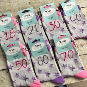 Peut inclure: Ensemble de six paires de chaussettes en coton blanc avec des étoiles violettes et roses. Chaque chaussette présente un numéro différent imprimé en rose : 18, 21, 30, 40, 50, 60 et 70.
