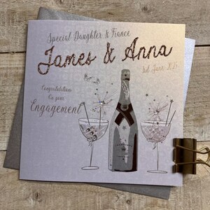 K&ouml;nnte beinhalten: Eine silberne und wei&szlig;e Verlobungskarte mit einer Champagnerflasche und zwei Gl&auml;sern. Die Karte lautet "Special Daughter & Fiance James & Anna 3rd June 2025 Congratulations on your Engagement".