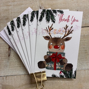 May include: A stack of white greeting cards featuring a cartoon reindeer with a red nose, holding a gift with a red bow. The reindeer has string lights on its antlers and the gift. The text Thank You is printed in red. Pine branches frame the top and bottom.