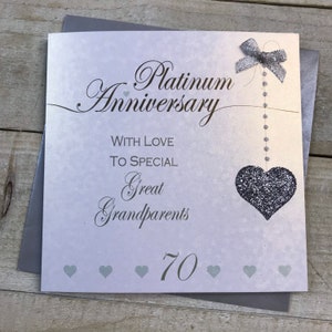 Puede incluir: Una tarjeta plateada y blanca con el texto "Platinum Anniversary" y "With Love To Special Great Grandparents 70". Un coraz&oacute;n de purpurina plateada cuelga de una cinta plateada.