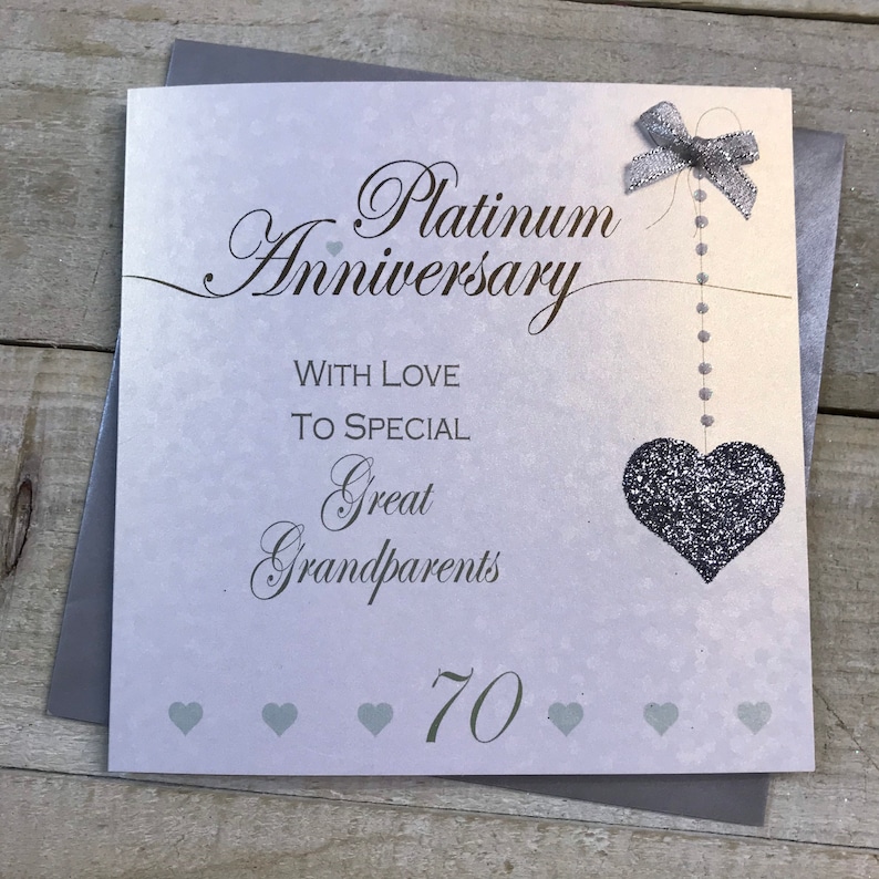 Puede incluir: Una tarjeta plateada y blanca con el texto "Platinum Anniversary" y "With Love To Special Great Grandparents 70". Un coraz&oacute;n de purpurina plateada cuelga de una cinta plateada.