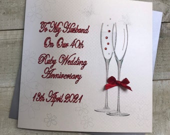 May include: A white card with red glitter text that reads "To My Husband On Our 40th Ruby Wedding Anniversary 13th April 2021". The card features a drawing of two champagne glasses with a red bow.