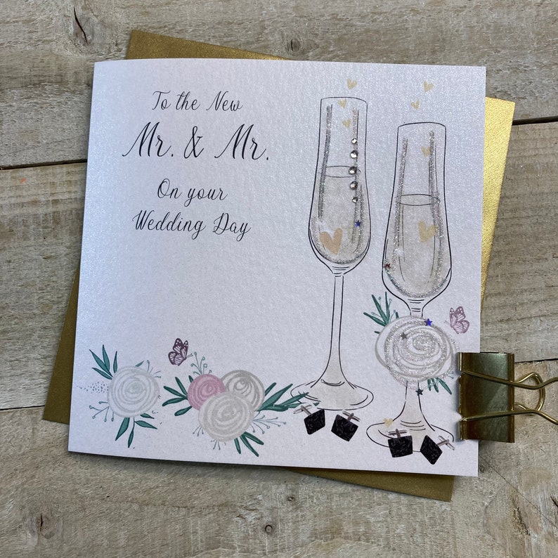 K&ouml;nnte beinhalten: Eine wei&szlig;e Karte mit goldfarbenem Rand und dem Text "To the New Mr. & Mr. On your Wedding Day" in schwarzer Schrift. Zwei Champagnergl&auml;ser mit goldenem Glitzer und Herzen sind auf die Karte gezeichnet. Am unteren Rand der Karte befindet sich ein florales Design mit wei&szlig;en und rosa Bl&uuml;ten und Schmetterlingen.