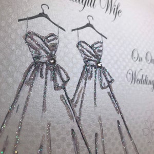 May include: A silver glitter card with two white wedding dresses on hangers. The card says "To Our Wonderful Wife" and "On Our Wedding Day".