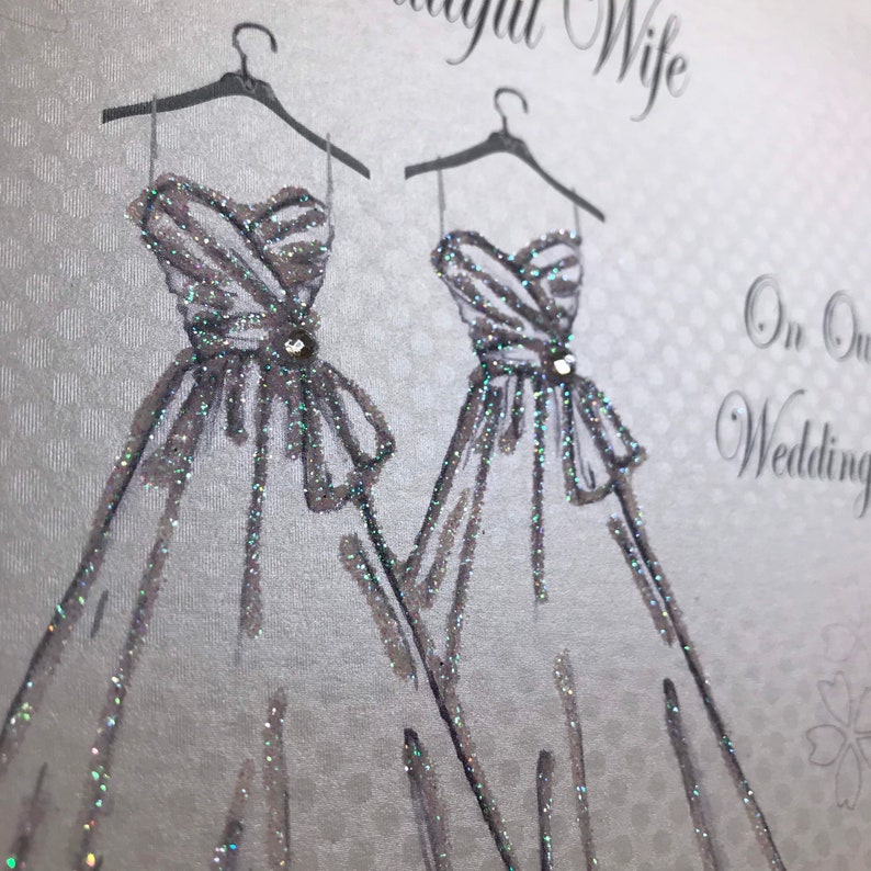 May include: A silver glitter card with two white wedding dresses on hangers. The card says "To Our Wonderful Wife" and "On Our Wedding Day".